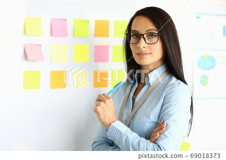 Businesswoman in blue shirt and glasses stand near blackboard with pen in her hand and smile. Businesswoman in blue shirt and glasses stand near blackboard with pen in her hand and smile. 69018373