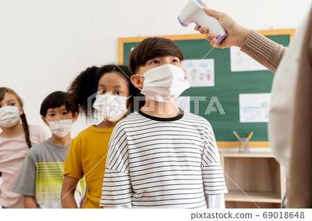 Group of diverse students in school building checked and scanned for temperature check. Elementary pupils are wearing a face mask and line up before entering into classroom. Group of diverse students in school building checked and scanned for temperature check. Elementary pupils are wearing a face mask and line up before entering into classroom. 69018648