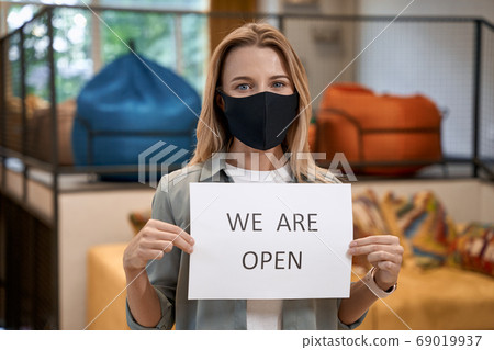 Young caucasian woman, female office worker in protective face mask showing paper with text WE ARE OPEN at camera while standing in the modern office 69019937