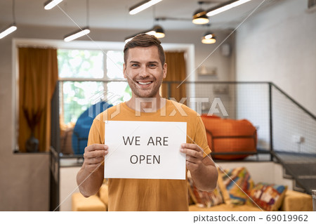 Opening office after lockdown. Young happy man showing paper with text WE ARE OPEN at camera and smiling while standing in the modern coworking space 69019962