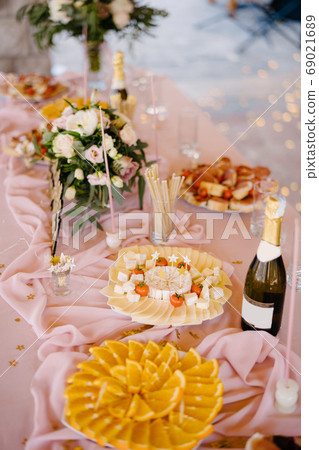 Wedding table decorated in pink flowers with sliced orange slices, cheese plate and sandwiches near a bouquet of flowers and a bottle of champagne. 69021689