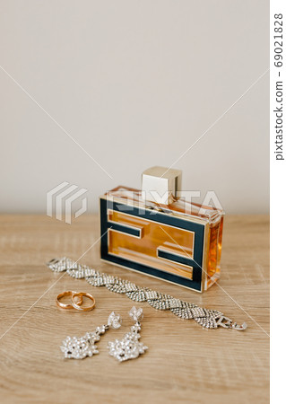 A perfume bottle, a bracelet, earrings-studs and wedding rings on a wooden table against a light wall. A perfume bottle, a bracelet, earrings-studs and wedding rings on a wooden table against a light wall. 69021828