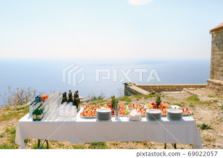 A festively served buffet table with water, champagne and fruits on plates against the backdrop of an old building by the water. A festively served buffet table with water, champagne and fruits on plates against the backdrop of an old building by the water. 69022057