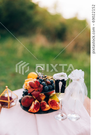 A painted table with a bookcase with fruits and glasses for champagne in the suits of the bride and groom. A painted table with a bookcase with fruits and glasses for champagne in the suits of the bride and groom. 69022152
