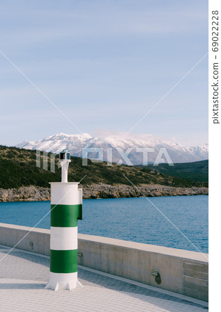 A small striped green and white lighthouse against the backdrop of the Adriatic Sea and the snow-capped peak of Mount Lovcen. 69022228