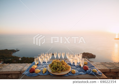 A table set for a celebration with clean empty glasses for champagne, a plate with white grapes and fruits on a blue cloth against a background of water. A table set for a celebration with clean empty glasses for champagne, a plate with white grapes and fruits on a blue cloth against a background of water. 69022354
