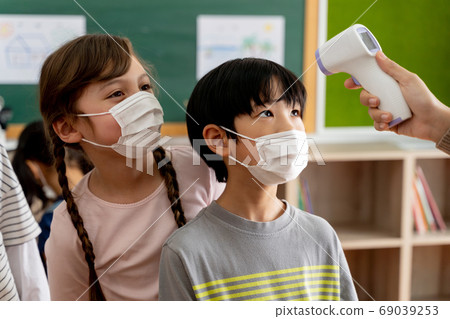 Group of diverse students in school building checked and scanned for temperature check. Elementary pupils are wearing a face mask and line up before entering into classroom. Group of diverse students in school building checked and scanned for temperature check. Elementary pupils are wearing a face mask and line up before entering into classroom. 69039253