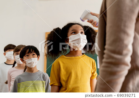 Group of diverse students in school building checked and scanned for temperature check. Elementary pupils are wearing a face mask and line up before entering into classroom. Group of diverse students in school building checked and scanned for temperature check. Elementary pupils are wearing a face mask and line up before entering into classroom. 69039271