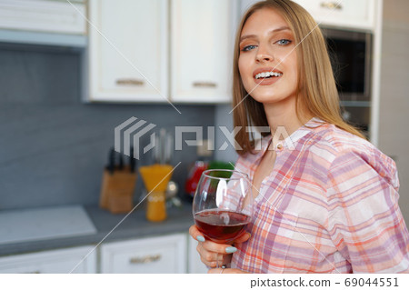 Blonde caucasian woman with glass of red wine standing in her kitchen Blonde caucasian woman with glass of red wine standing in her kitchen 69044551