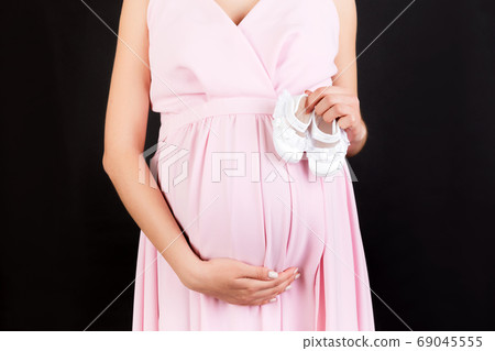 Cropped image of pregnant woman in pink dress holding baby girl booties against her belly at black background. Happy pregnancy. Copy space Cropped image of pregnant woman in pink dress holding baby girl booties against her belly at black background. Happy pregnancy. Copy space 69045555