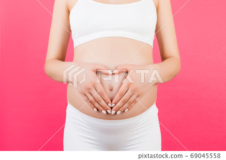 Cropped image of pregnant woman in white underwear making heart shape with the hands on her abdomen at pink background. Baby bump concept. Copy space Cropped image of pregnant woman in white underwear making heart shape with the hands on her abdomen at pink background. Baby bump concept. Copy space 69045558