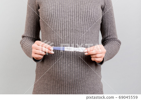 Cropped image of positive pregnancy test with two stripes against pregnant woman's abdomen at gray background. Future mother in gray dress. Pregnancy surprise. Copy space 69045559