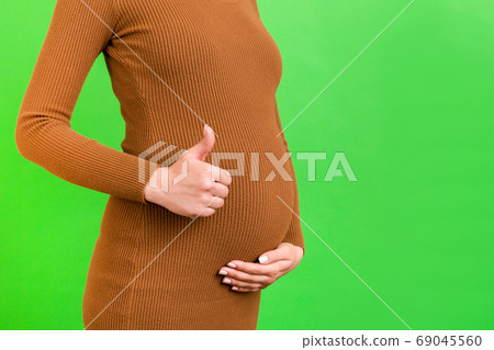 Cropped shot of positive pregnant woman in brown dress showing thumb up cool gesture over her baby bump at green background. Happy maternity. Copy space Cropped shot of positive pregnant woman in brown dress showing thumb up cool gesture over her baby bump at green background. Happy maternity. Copy space 69045560