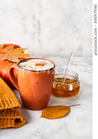 Pumpkins spice latte or coffee with creamy foam in orange cup with pumpkin, leaves, jar of honey and white cozy sweater on white 69047698