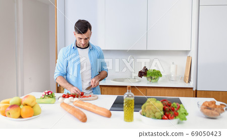Handsome man cuts tomatoes on a wooden board to preparing salad from fresh vegetables at cozy kitchen. Healthy food. Handsome man cuts tomatoes on a wooden board to preparing salad from fresh vegetables at cozy kitchen. Healthy food. 69050423