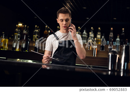 the bartender is talking on the phone at the bar against the background of bottles with a bar spoon in his hands 69052883