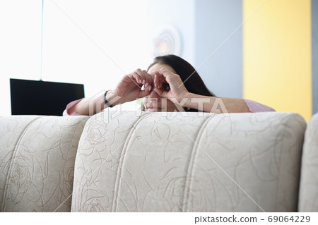 Woman looks out from behind sofa and holds her hands through binoculars. Woman looks out from behind sofa and holds her hands through binoculars. 69064229
