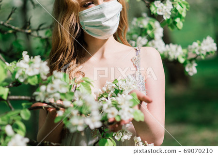 Beautiful young bride in a wedding dress and a white medical mask on her face near a blooming apple. Beautiful young bride in a wedding dress and a white medical mask on her face near a blooming apple. 69070210