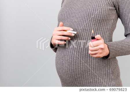 Close up of a cigarette and a lighter in pregnant woman's hands at gray background. Unhealthy habit during pregnancy. Smoking abuse Close up of a cigarette and a lighter in pregnant woman's hands at gray background. Unhealthy habit during pregnancy. Smoking abuse 69079823