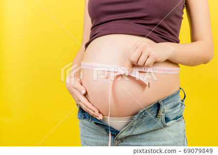 Close up of a pregnant woman in opened jeans measuring her belly with a tape measure at colorful background with copy space. entimeter measurement Close up of a pregnant woman in opened jeans measuring her belly with a tape measure at colorful background with copy space. entimeter measurement 69079849