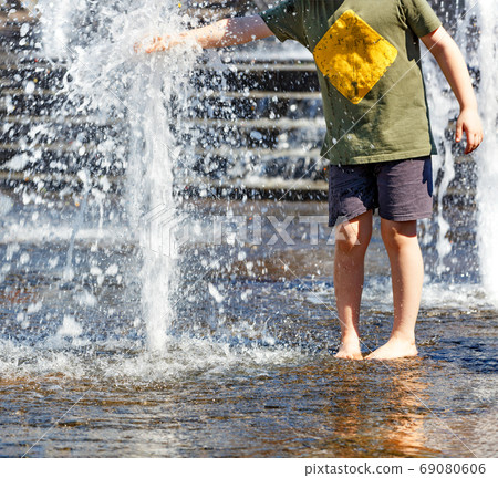 A teenager stands barefoot by a fountain and tries to stop a powerful stream of water with his hand on a bright hot day. 69080606
