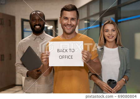 Young happy caucasian man, office worker showing paper with text BACK TO OFFICE at camera and smiling while standing with colleagues in the office Young happy caucasian man, office worker showing paper with text BACK TO OFFICE at camera and smiling while standing with colleagues in the office 69080804