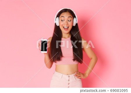 Technology, communication and online lifestyle concept. Excited and happy asian girl looking amazed, showing smartphone screen while listening podcast or music in headphones, pink background Technology, communication and online lifestyle concept. Excited and happy asian girl looking amazed, showing smartphone screen while listening podcast or music in headphones, pink background 69081095