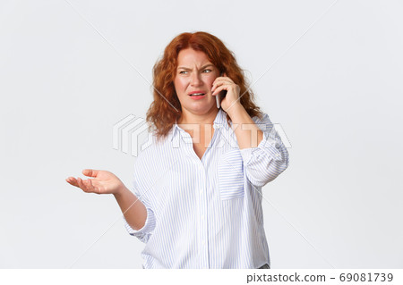 Frustrated and displeased, confused redhead middle-aged woman shrugging and grimacing disgusted while talking on mobile phone about something awful, standing white background Frustrated and displeased, confused redhead middle-aged woman shrugging and grimacing disgusted while talking on mobile phone about something awful, standing white background 69081739