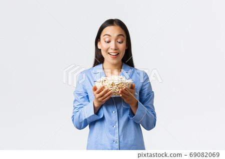 Home leisure, sleepover and slumber party concept. Excited smiling asian girl in pajamas looking tempting at tasty bowl of popcorn, prepared snacks for movie night, white background 69082069