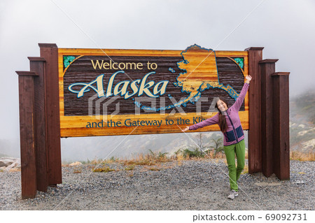 Alaska border sign tourist posing at welcome billboard - Happy Asian woman traveling in USA. Alaska border sign tourist posing at welcome billboard - Happy Asian woman traveling in USA. 69092731