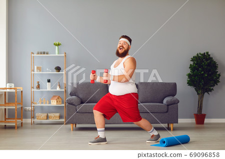 Funny fat man in red shorts and white t-shirt smiling doing exercises in the room Funny fat man in red shorts and white t-shirt smiling doing exercises in the room 69096858