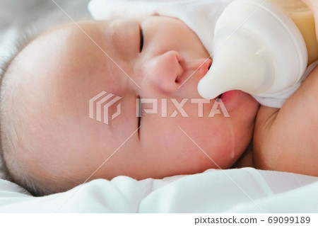 Portrait of a newborn Asian baby on the bed drinks milk from a bottle, Charming black-eyed baby 4 month old lies in bed ,A child resting on a bed 69099189