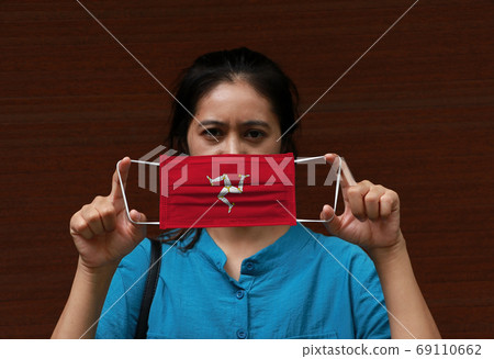 A woman and hygienic mask with Isle of man flag pattern in her hand and raises it to cover her face 69110662