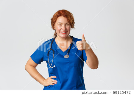 Medicine, healthcare and coronavirus concept. Professional doctor, female medical worker in blue scrubs ensure all good, showing thumb-up and nod in approval, compliment choice, grey background 69115472