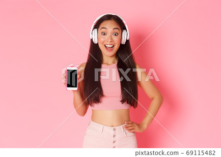 Technology, communication and online lifestyle concept. Excited and happy asian girl looking amazed, showing smartphone screen while listening podcast or music in headphones, pink background 69115482