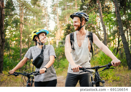 Cheerful couple pushing bicycles and walking along the forest road. Happy couple with bicycle walking through park and talking and laugh. Caucasian young man and woman riding bicycles in woods 69116654