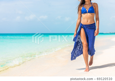 Woman walking on beach in blue fashion beachwear bathing suit and sarong pareo sun skirt relaxing in luxury Caribbean vacation holidays. Summer or winter getaway destination. 69120510
