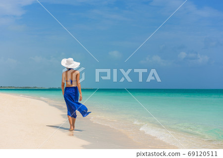 Luxury travel woman walking on perfect beach vacation Caribbean destination in blue dress skirt sarong. Elegant lady tourist relaxing on summer vacations wearing sun hat. Luxury travel woman walking on perfect beach vacation Caribbean destination in blue dress skirt sarong. Elegant lady tourist relaxing on summer vacations wearing sun hat. 69120511