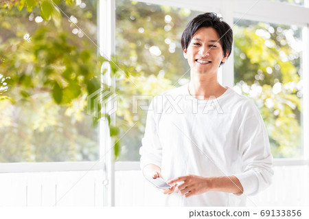 A man wearing a white sewn standing by the window with a smartphone in hand 69133856