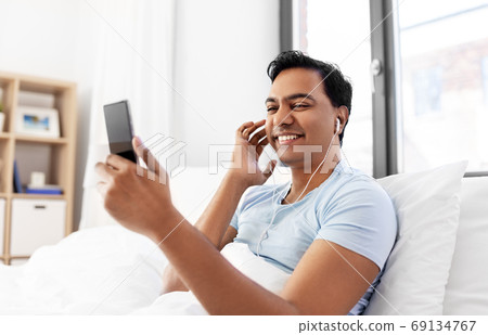 indian man in earphones with phone in bed at home indian man in earphones with phone in bed at home 69134767