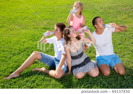 Parents with teenager son and daughter drinking cold water from plastic bottles in summer park Parents with teenager son and daughter drinking cold water from plastic bottles in summer park 69137830