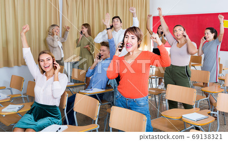 Group of adult students expressing happiness while talking on phones after passing exam in class 69138321