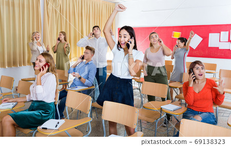 Group of adult students expressing happiness while talking on phones after passing exam in class 69138323