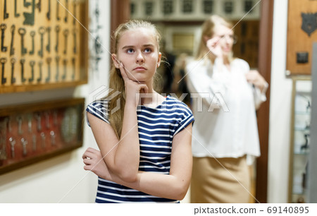 Upset teen girl standing in museum of art, mother on background Upset teen girl standing in museum of art, mother on background 69140895