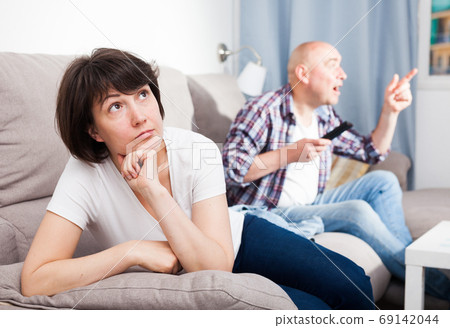 Sad woman sitting on the sofa. Next to her husband watching TV Sad woman sitting on the sofa. Next to her husband watching TV 69142044