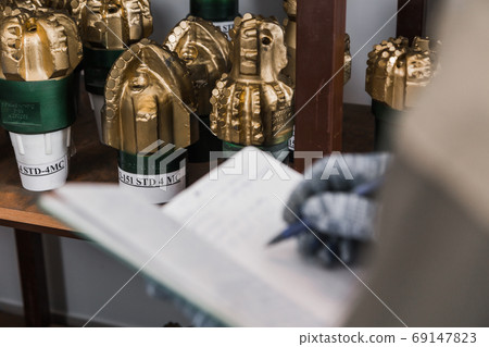 Engineer keeps records of drill bits in notebook at warehouse of production factory. Brand new PDC Polycrystalline Diamond Compact drilling bit on shelving in stock 69147823