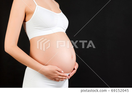 Cropped image of pregnant woman in white underwear embracing her big abdomen at black background. Maternity concept. Copy space Cropped image of pregnant woman in white underwear embracing her big abdomen at black background. Maternity concept. Copy space 69152045