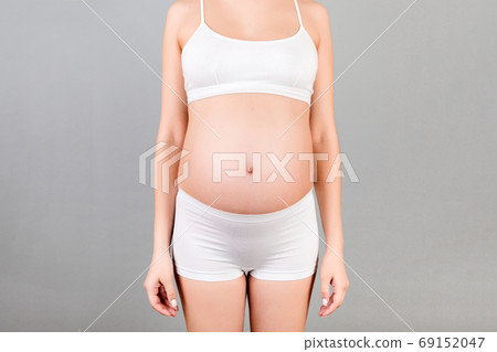 Cropped shot of pregnant abdomen in white underwear at gray background. Young woman is waiting for a baby. Copy space Cropped shot of pregnant abdomen in white underwear at gray background. Young woman is waiting for a baby. Copy space 69152047