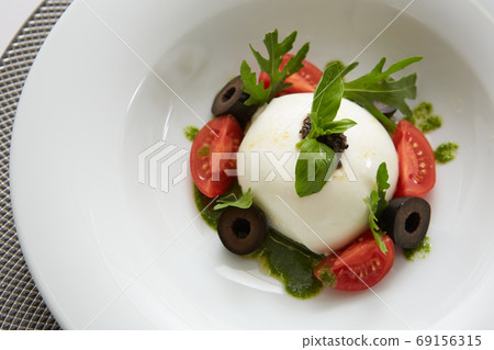 Traditional authentic soft and creamy fresh Italian burrata cheese shaped as white round ball over Italian tomatoes topped with rucola along side pesto reduction Traditional authentic soft and creamy fresh Italian burrata cheese shaped as white round ball over Italian tomatoes topped with rucola along side pesto reduction 69156315
