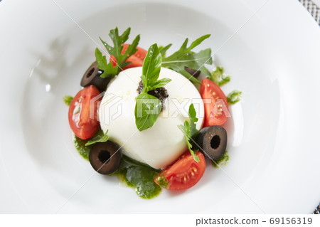 Traditional authentic soft and creamy fresh Italian burrata cheese shaped as white round ball over Italian tomatoes topped with rucola along side pesto reduction Traditional authentic soft and creamy fresh Italian burrata cheese shaped as white round ball over Italian tomatoes topped with rucola along side pesto reduction 69156319